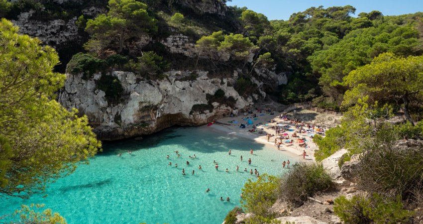 Vista aérea de Cala Macarelleta, con aguas turquesas y familias disfrutando de un día de playa entre los acantilados