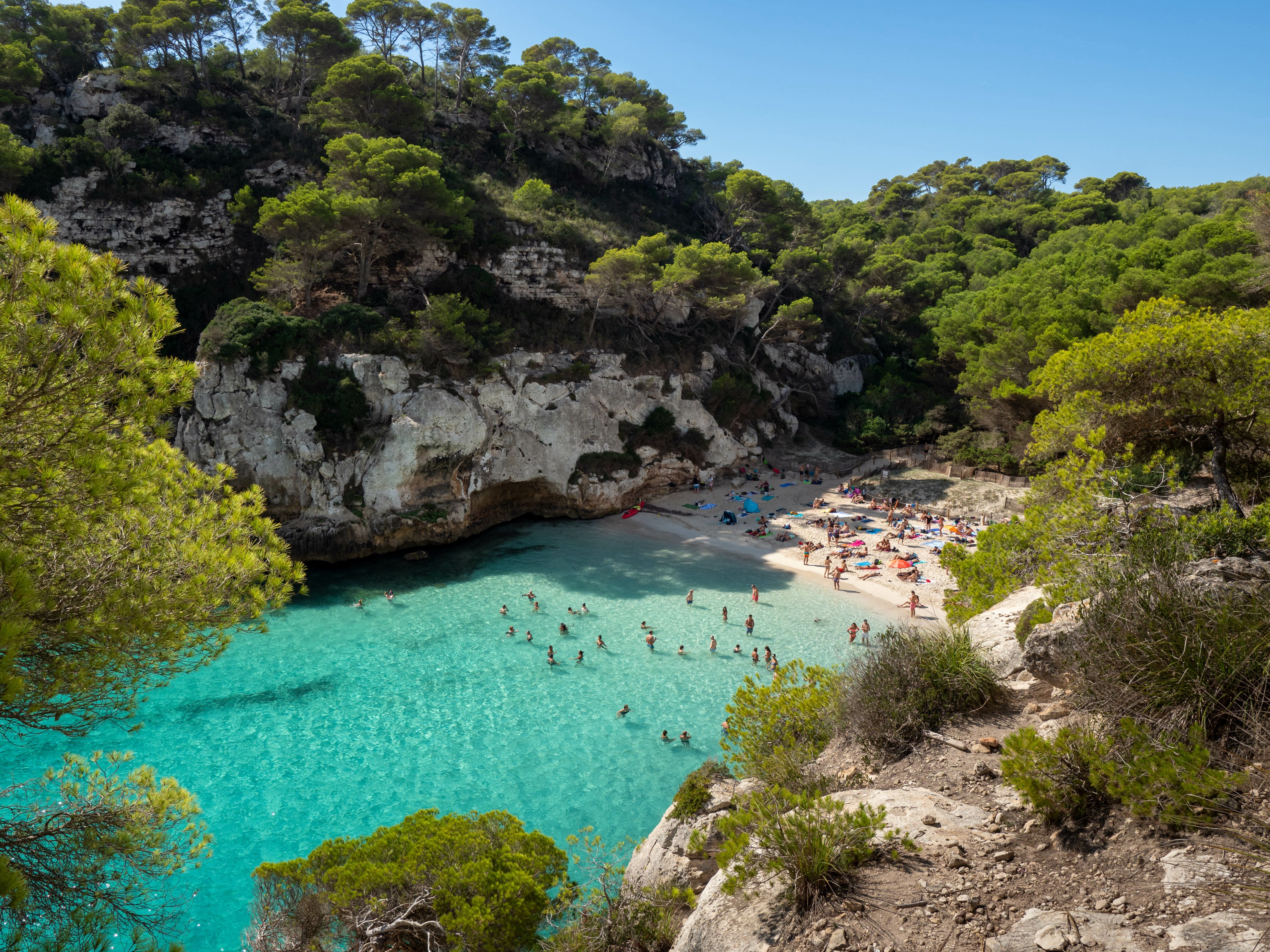 Aerial view of Cala Macarelleta, with turquoise waters and families enjoying a day at the beach among the cliffs.