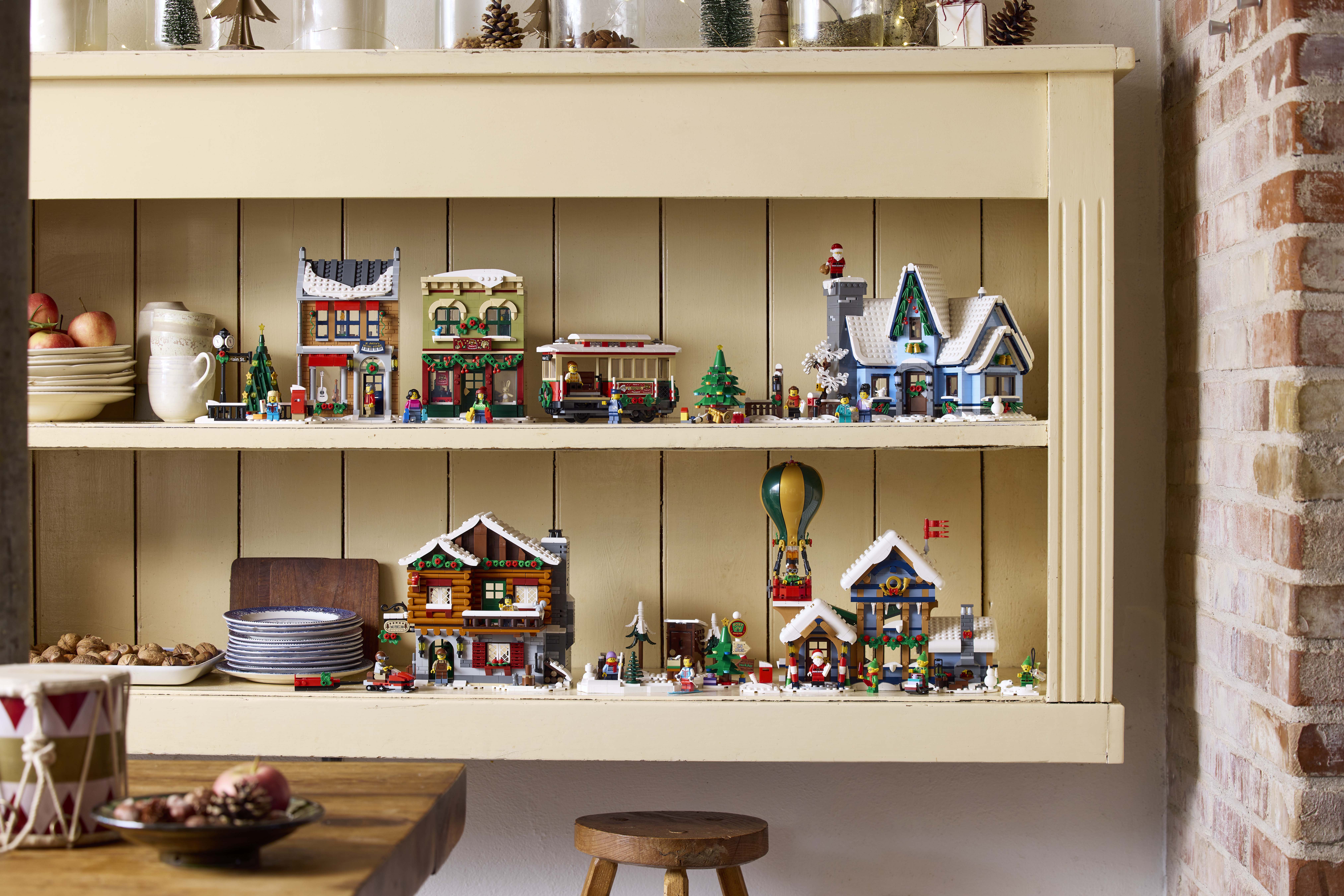 Consigue hasta 10 Avios por cada 1€ que gastes con Lego. En la imagen, las construcciones de Lego de la colección de Navidad sobre una estantería de madera, rodeadas por platos y vasos de temática navideña, con manzanas y galletas recién horneadas 