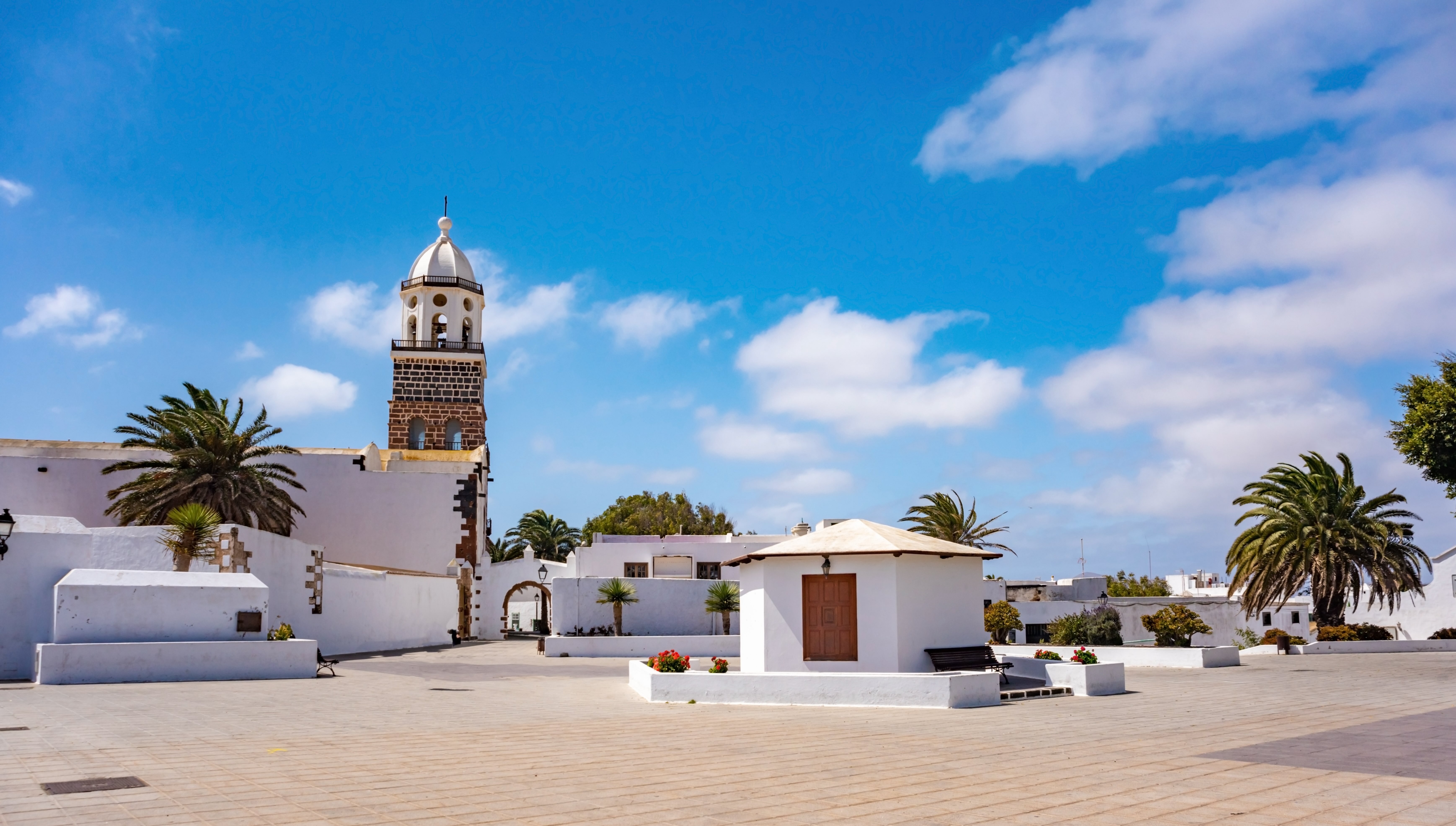 The picture shows one of the squares in Teguise, with whitewashed buildings and houses next to green palm trees.