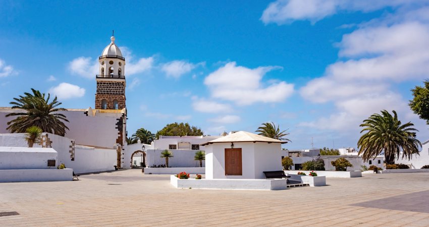 The picture shows one of the squares in Teguise, with whitewashed buildings and houses next to green palm trees.