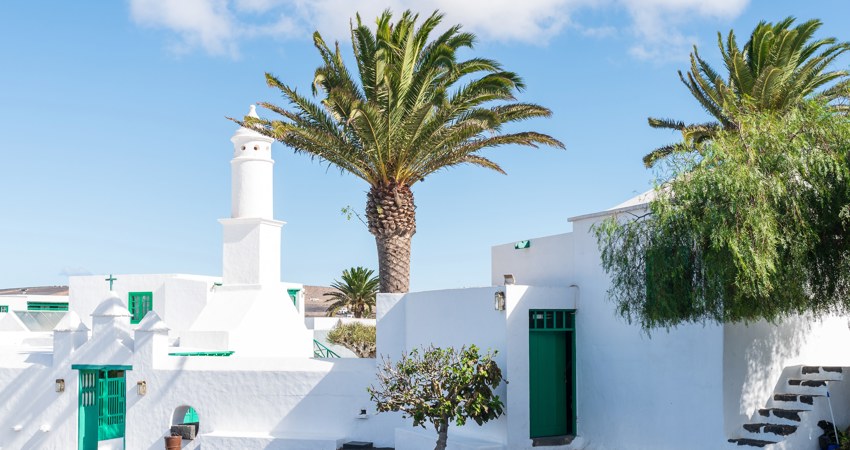 In the picture, one of the squares of San Bartolomé, with low houses dotted with palm trees. In the background is the profile of César Manrique's Monument to Fertility