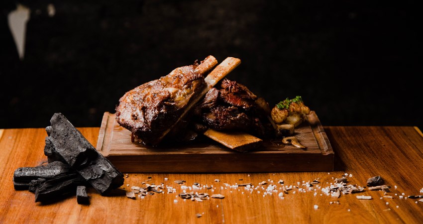 Presentation of a grilled rack of ribs at La Carnicería, on a wooden board, accompanied by a still life of salt and charcoal.