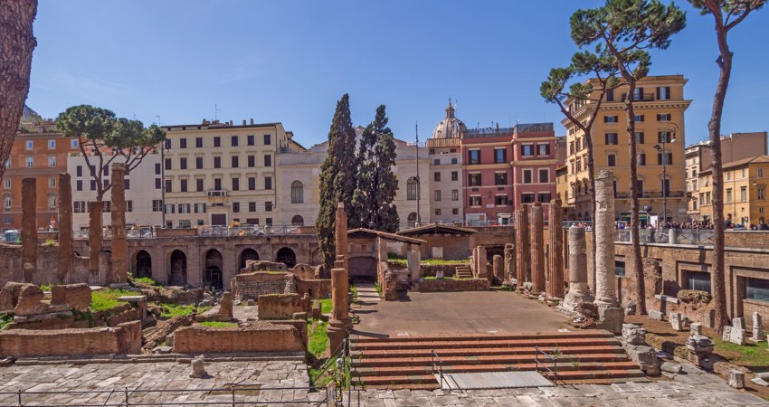 En la imagen,Largo di Torre Argentina, con los escalones donde César fue asesinado en la parte derecha de la panorámica y columnas, arcadas y vegetación en la parte izquierda. Los edificios de Roma enmarcan el paisaje 