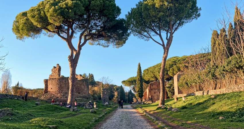 View of the tessellated roadway of the Appian Way, surrounded by nature, with trees and archaeological remains on either side of the road.
