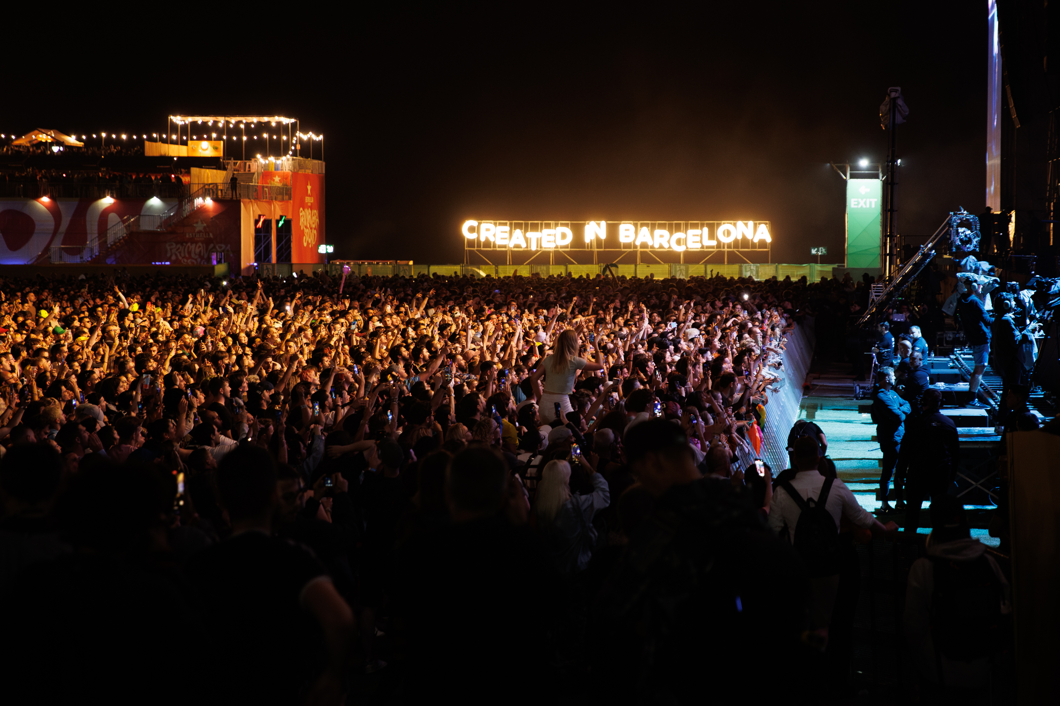 Primavera Sound 2025. In the image, dark night with thousands of people dancing and singing in front of a stage. In the background, a sign with yellow neon lights where you that reads “Created in Barcelona”.
