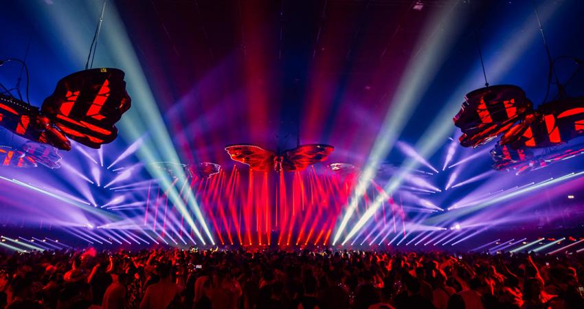 En la imagen, mariposas gigantes vuelan sobre la multitud frente al escenario, perseguidas por columnas de luz
