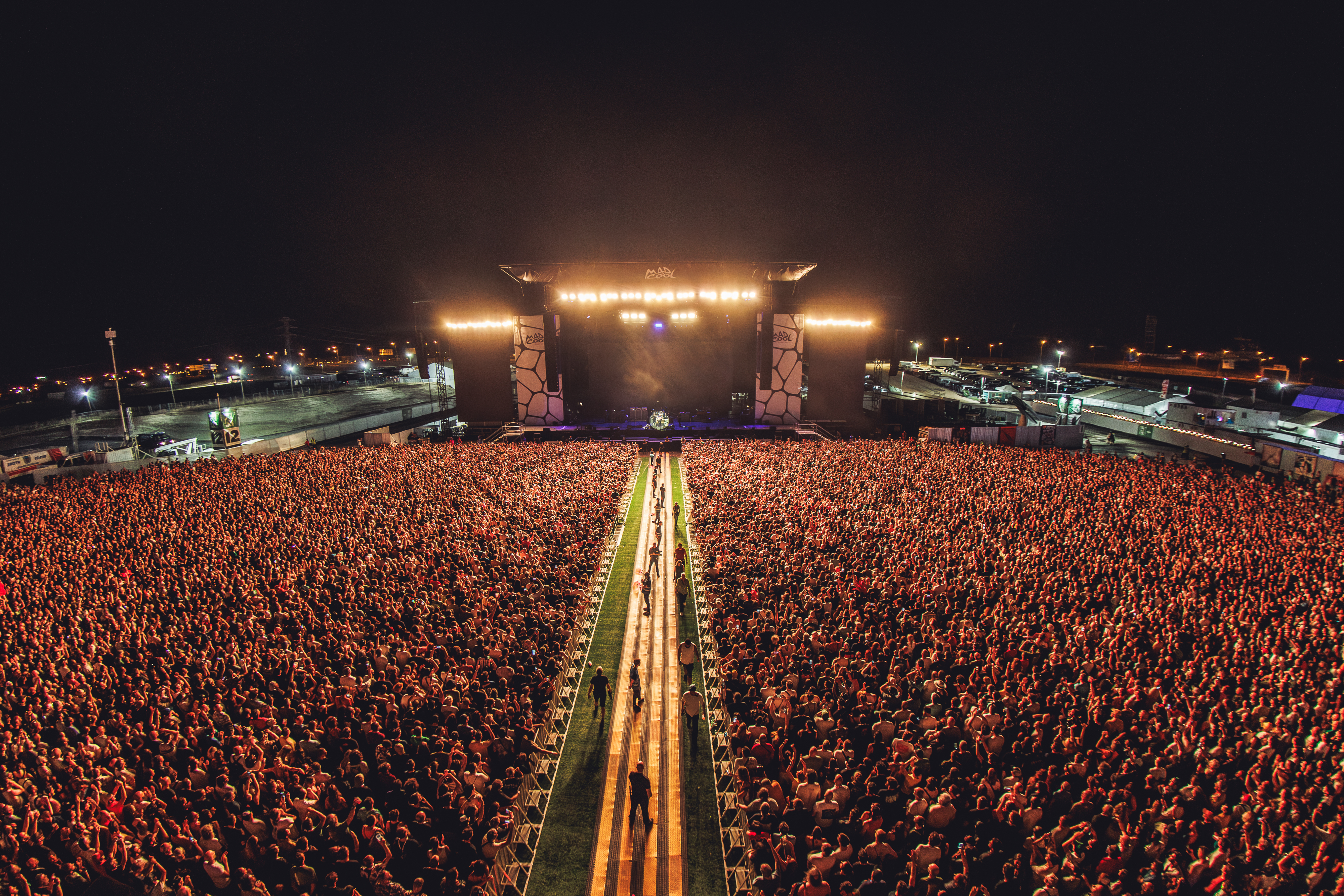 The image shows an aerial view of the main stage at Mad Cool's last edition. The stage is the focal point of the photo, with a path dividing a crowd of people that spills over the edges of the image.