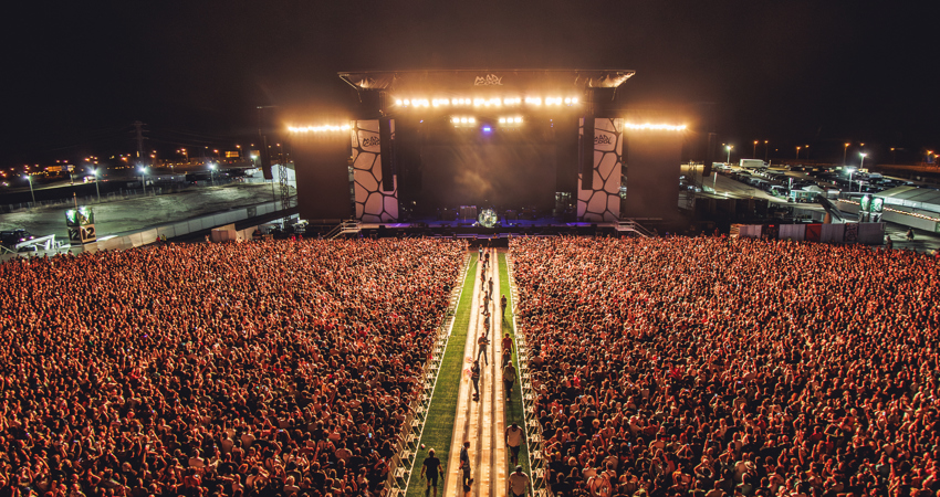 The image shows an aerial view of the main stage at Mad Cool's last edition. The stage is the focal point of the photo, with a path dividing a crowd of people that spills over the edges of the image.