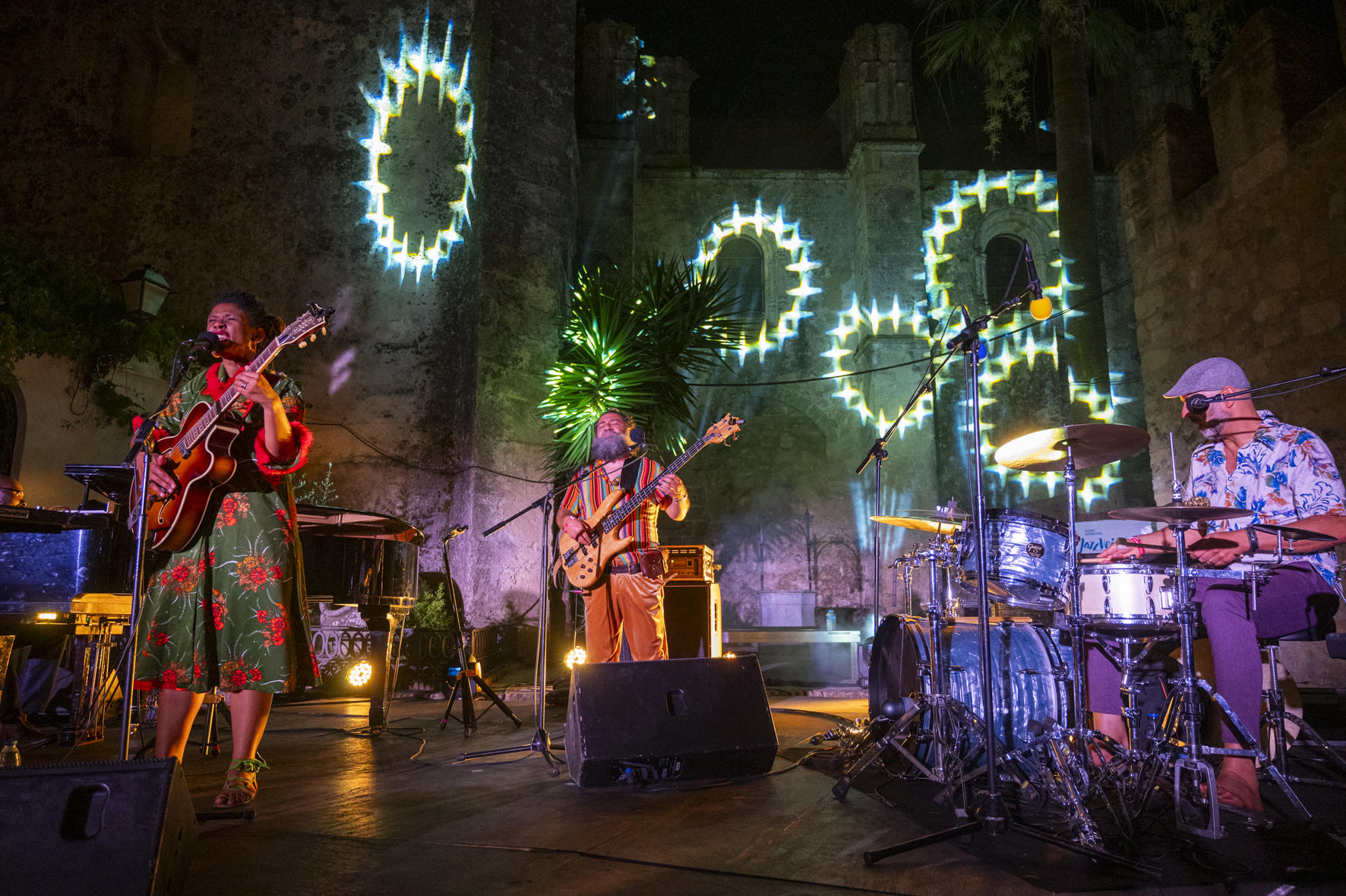 In the image, three artists are playing the bass, guitar and drums, respectively. In the background, images are projected onto the wall.