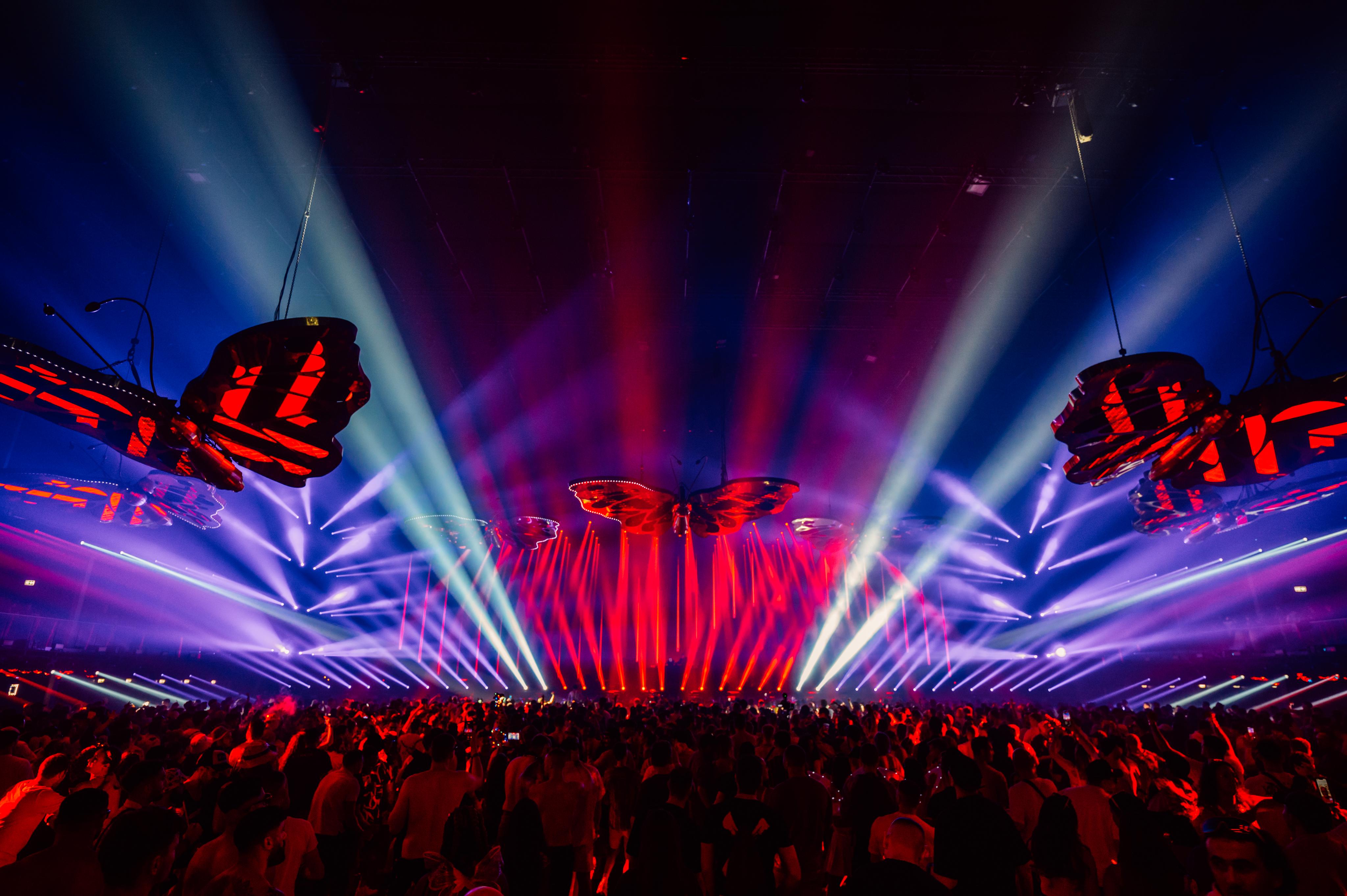 The image shows giant butterflies flying over the crowd in front of the stage, pursued by columns of light.