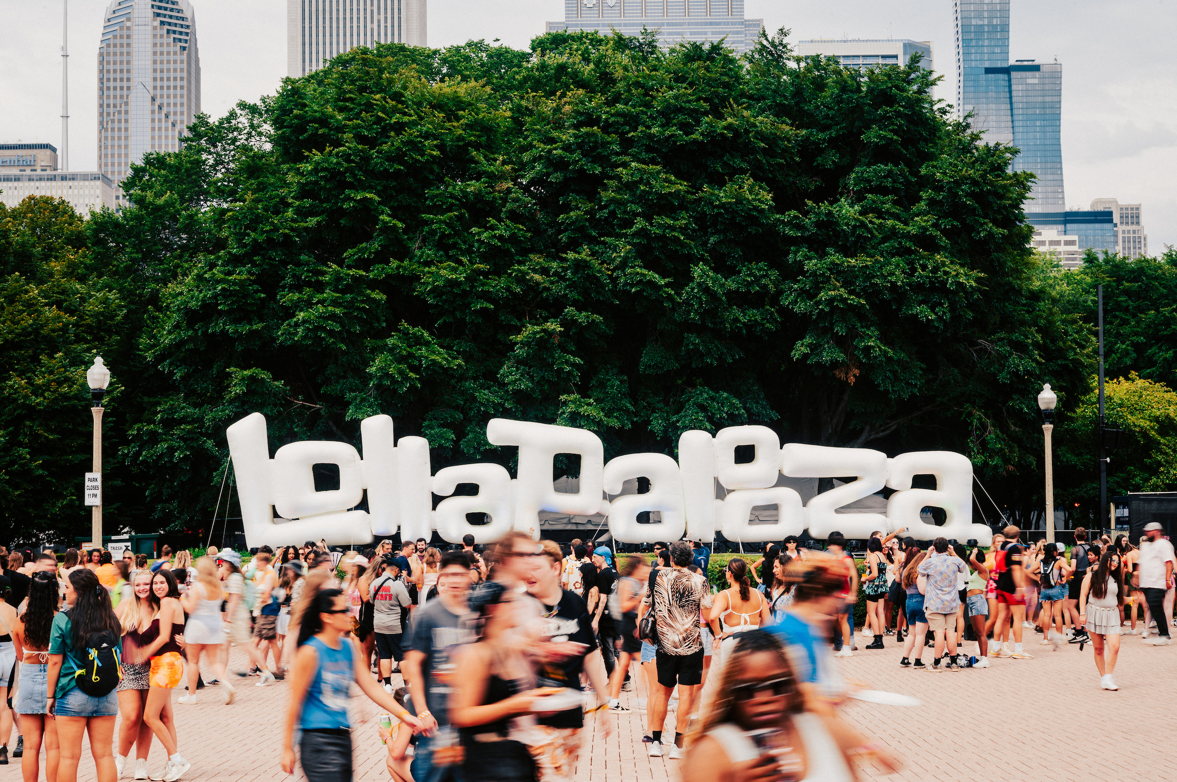 The image shows giant white letters spelling out 'Lollapalooza'. There is vegetation in the background and visitors around.