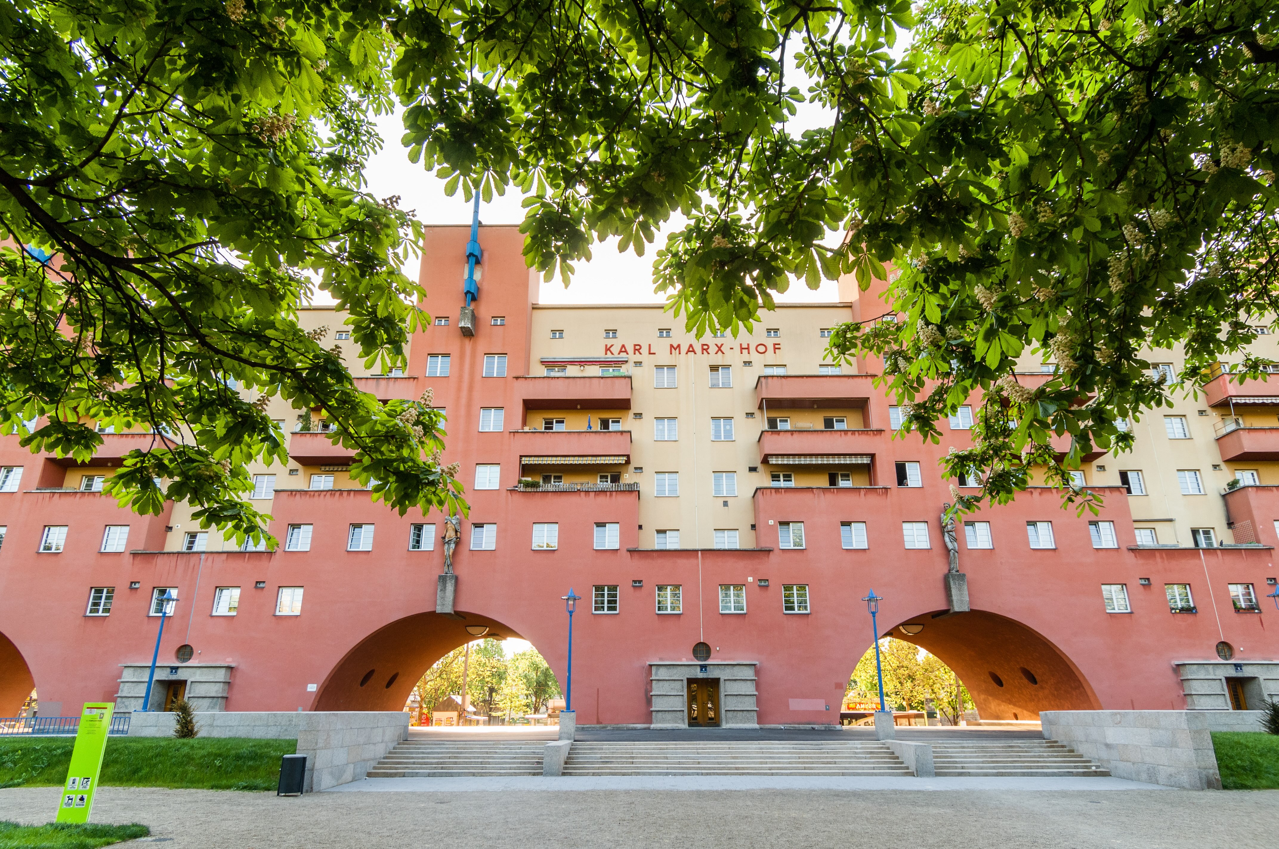 In the image, one of the entrances to Karl-Marx-Hof, with trees surrounding the building. The archways leading to the inner courtyards can be seen. Between them are the different entrances to the flats, in white and red buildings.