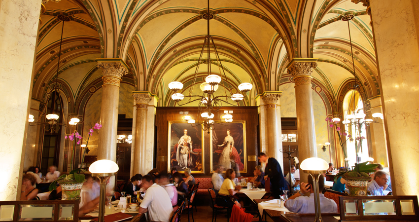 Café Central in Vienna. In the background, two portraits of Franz Joseph I of Austria and Empress Elisabeth (Sissi). Tables filled with customers enjoy the elegant atmosphere under classic hanging lamps.
