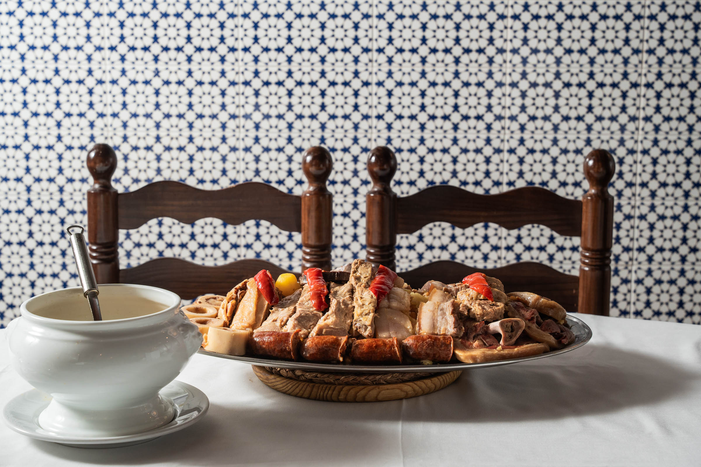 En primer plano, una mesa de mantel blanco con sopera y una gran bandeja de carnes. Frente a ellas dos sillas de taberna, de madera y mimbre y un fondo de azulejos azules y blancos con motivos geométricos 