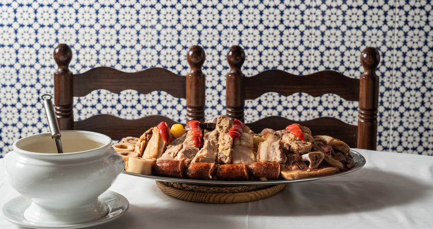 En primer plano, una mesa de mantel blanco con sopera y una gran bandeja de carnes. Frente a ellas dos sillas de taberna, de madera y mimbre y un fondo de azulejos azules y blancos con motivos geométricos 
