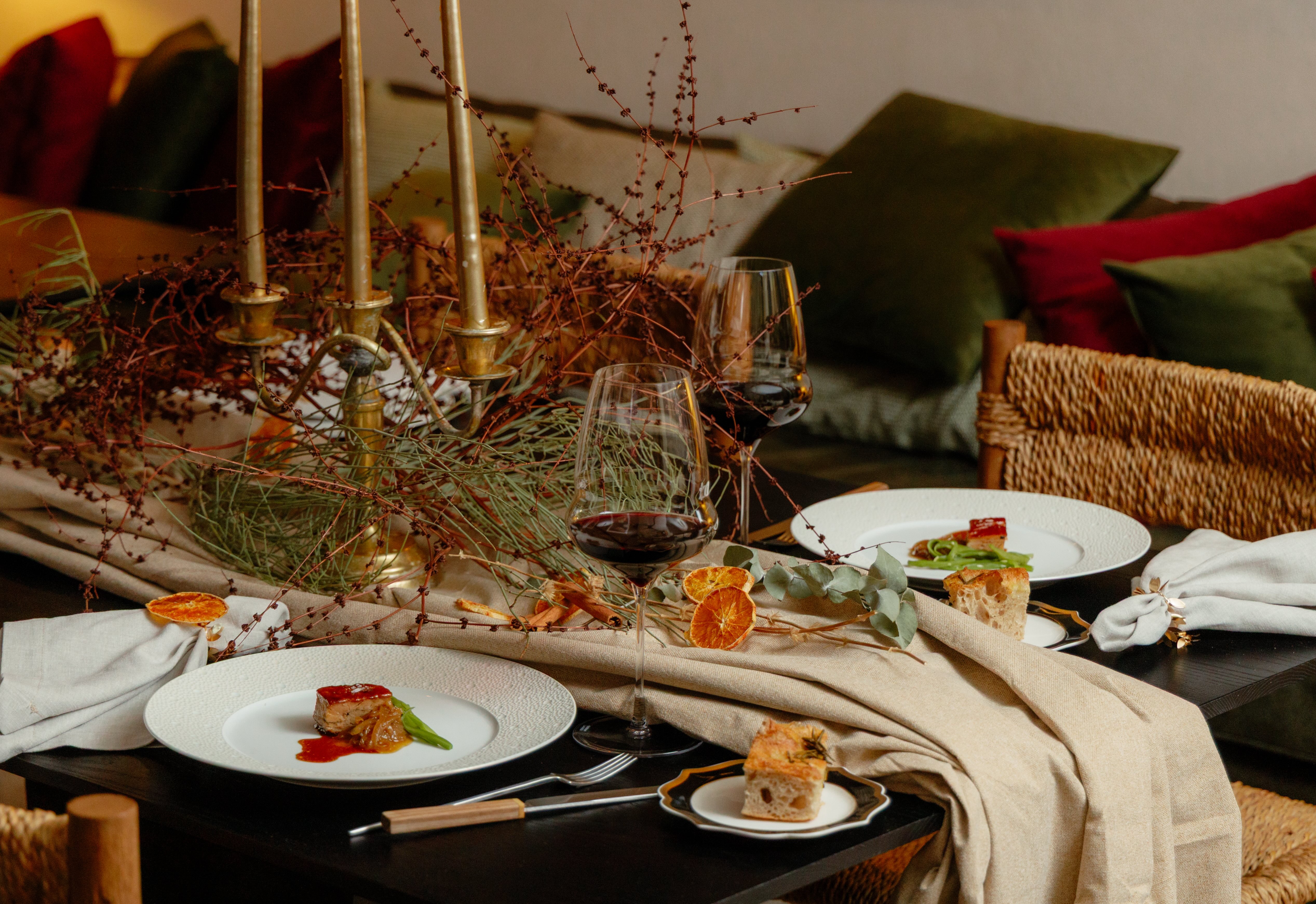 In the image, one of Gurisa's tables, with one of the main dishes from the Christmas menu, red wine and bread, and a table decoration made with fabrics, candlesticks, autumn plants and dried fruit.
