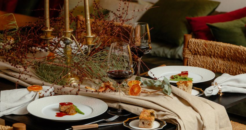 In the image, one of Gurisa's tables, with one of the main dishes from the Christmas menu, red wine and bread, and a table decoration made with fabrics, candlesticks, autumn plants and dried fruit.
