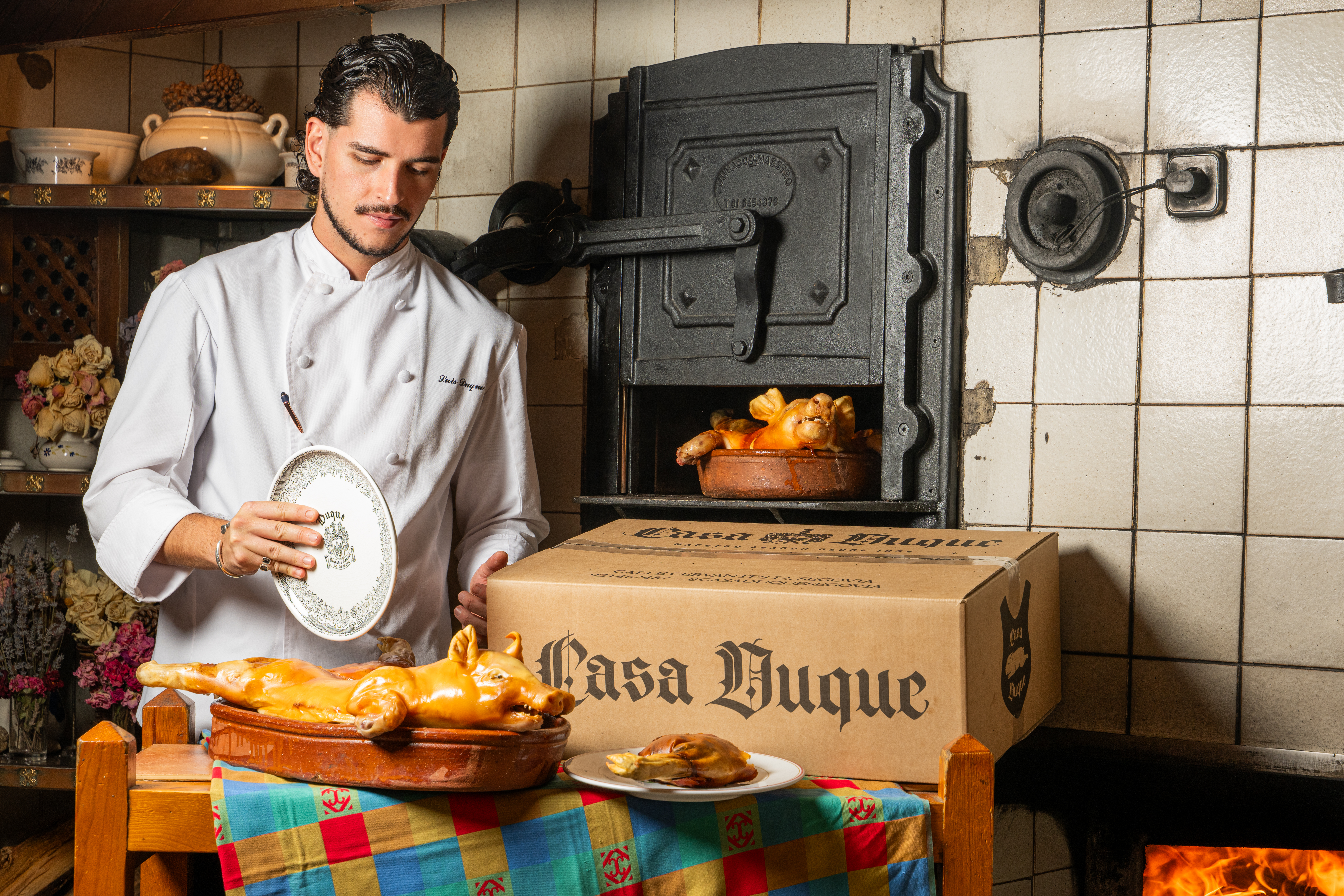 In the image, a chef from Casa Duque prepares to carve a freshly roasted suckling pig on a clay tray in the traditional manner (with a plate). Next to him is one of Casa Duque's food delivery boxes, and behind it, an iron oven where another piece is being cooked
