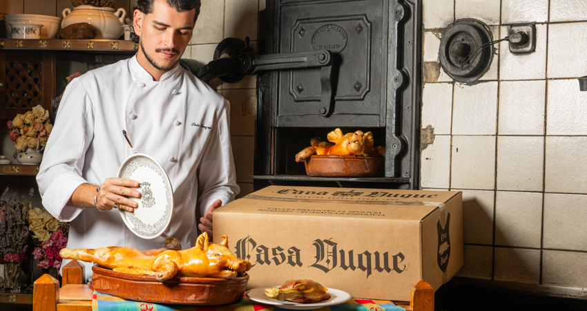 In the image, a chef from Casa Duque prepares to carve a freshly roasted suckling pig on a clay tray in the traditional manner (with a plate). Next to him is one of Casa Duque's food delivery boxes, and behind it, an iron oven where another piece is being cooked