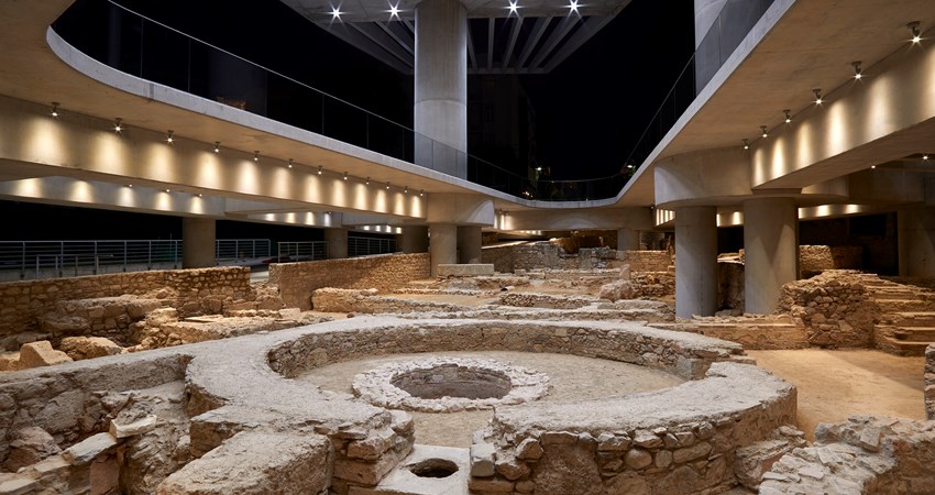 The image shows an archaeological excavation site with stone foundations and walls of varying heights and circular structures. Visitors can enjoy the space by walking along the suspended walkways.