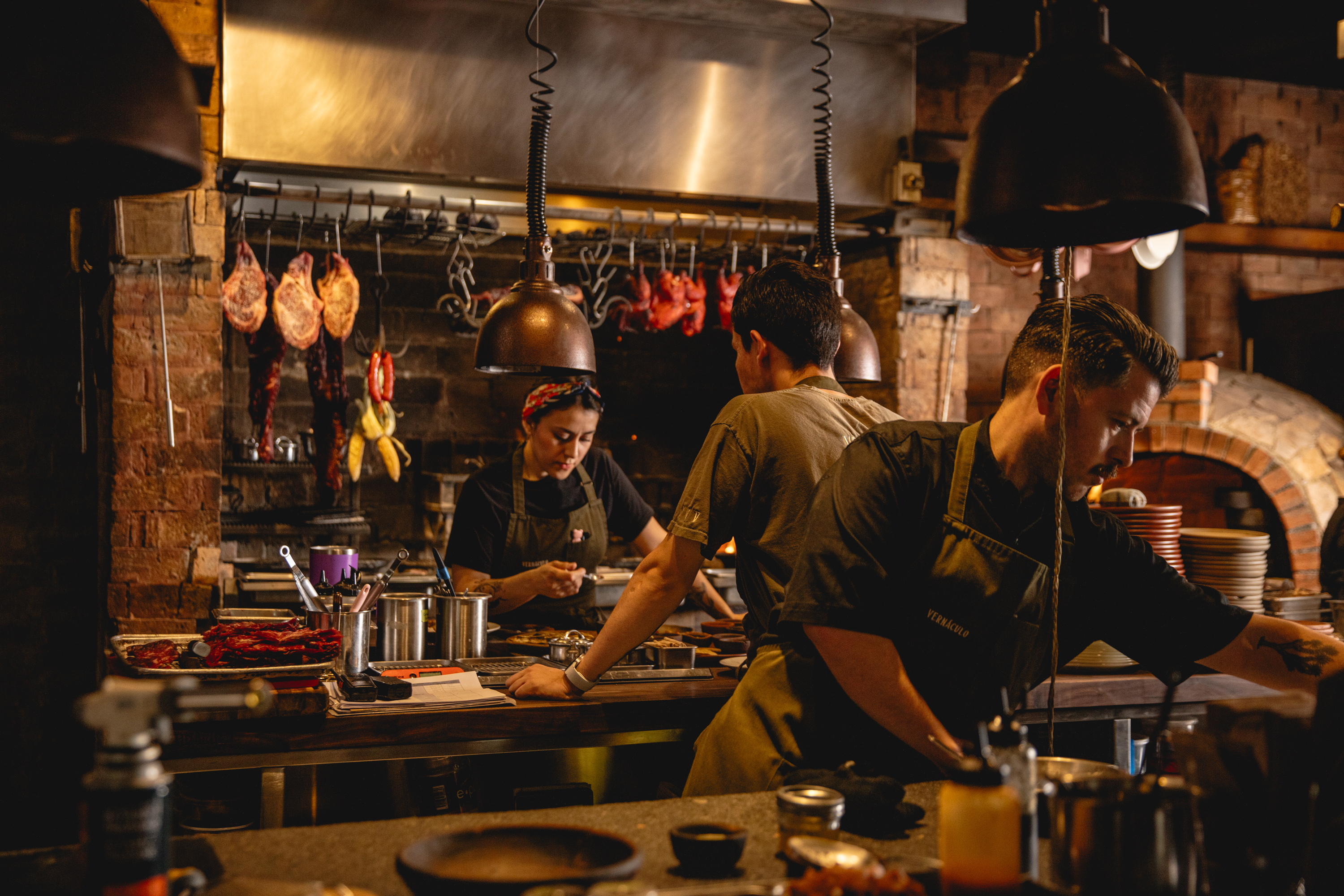 Vista del área de preparación en Vernáculo, con su estilo industrial. En primer plano, un chef de perfil organiza la mesa; al fondo, una mujer con pañuelo en la cabeza y otro cocinero trabajan bajo lámparas cálidas, rodeados de cortes de carne y pollos colgados sobre una parrilla de ladrillo.