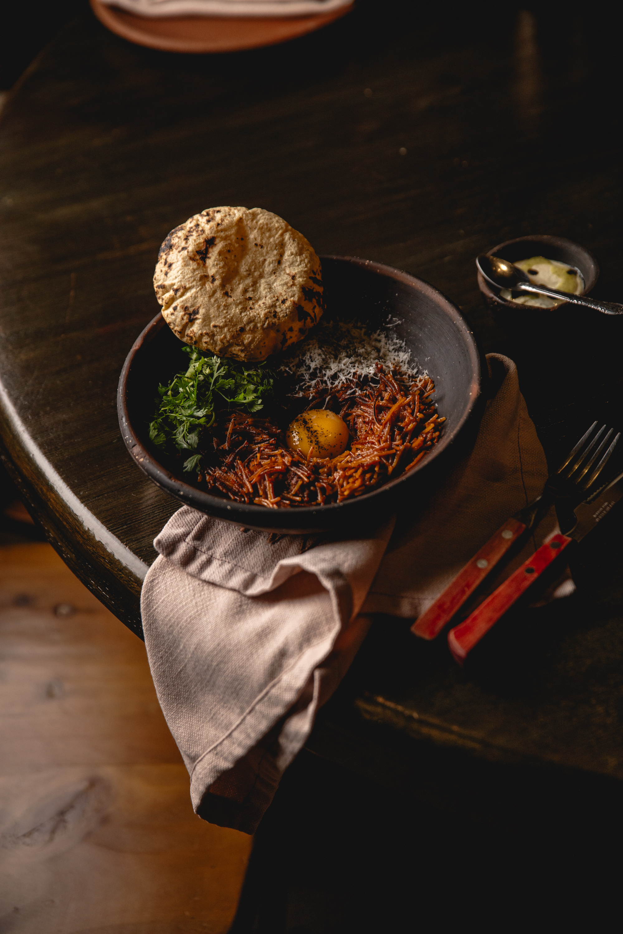Fotografía cenital de un cuenco de barro oscuro sobre una mesa de madera. El plato contiene fideos rojos tipo "fideo seco", coronados con una yema de huevo cruda, queso rallado, cilantro fresco y una tortilla de maíz tostada al fuego.